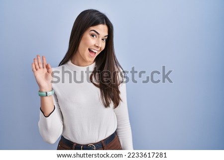 Young brunette woman standing over blue background waiving saying hello happy and smiling, friendly welcome gesture 
