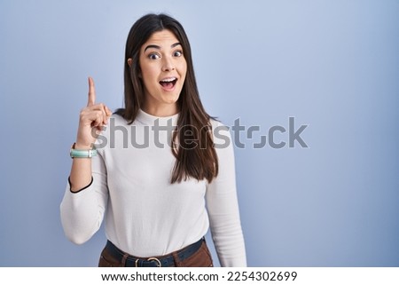 Young brunette woman standing over blue background pointing finger up with successful idea. exited and happy. number one. 