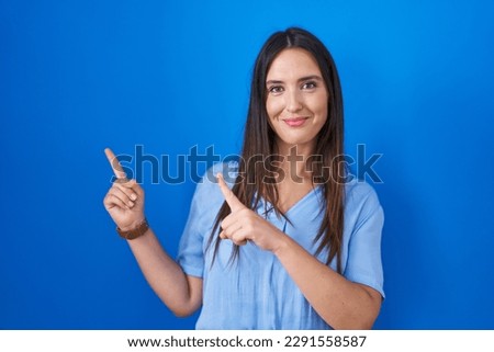 Young brunette woman standing over blue background smiling and looking at the camera pointing with two hands and fingers to the side. 