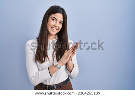 Young brunette woman standing over blue background clapping and applauding happy and joyful, smiling proud hands together 
