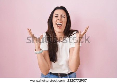 Young brunette woman standing over pink background crazy and mad shouting and yelling with aggressive expression and arms raised. frustration concept. 