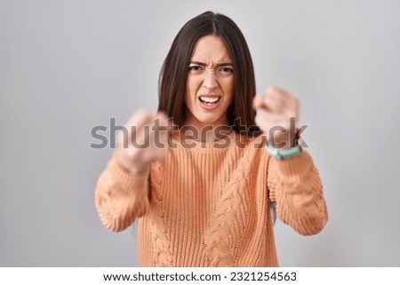 Young brunette woman standing over white background angry and mad raising fists frustrated and furious while shouting with anger. rage and aggressive concept. 