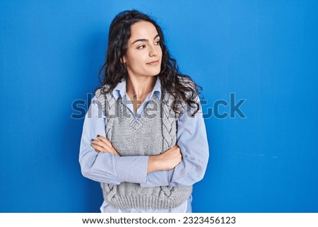 Young brunette woman standing over blue background looking to the side with arms crossed convinced and confident 