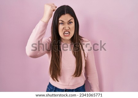 Young brunette woman standing over pink background angry and mad raising fist frustrated and furious while shouting with anger. rage and aggressive concept. 