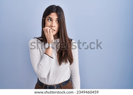 Young brunette woman standing over blue background looking stressed and nervous with hands on mouth biting nails. anxiety problem. 