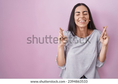 Young brunette woman standing over pink background gesturing finger crossed smiling with hope and eyes closed. luck and superstitious concept. 