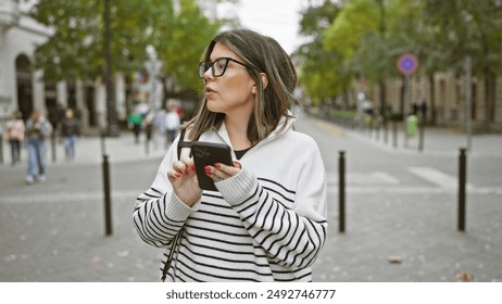 Young brunette woman in striped sweater using smartphone on european city street, surrounded by autumn foliage. - Powered by Shutterstock