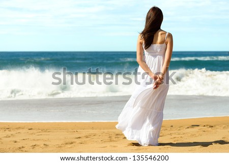 Young brunette woman in summer white dress standing on beach and looking to the sea. Caucasian girl relaxing and enjoying peace on vacation.