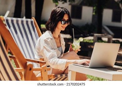 Young Brunette Woman in Sunglasses Working on Laptop and Drinking Cocktail While Sitting on Terrace of the Restaurant, Freelancer at Work - Powered by Shutterstock