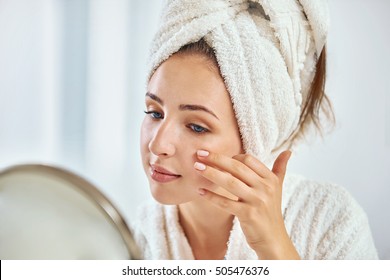 Eine junge Brunettenfrau mit einem Handtuch, das um den Kopf umhüllt ist und ihr Gesicht im runden Spiegel untersucht