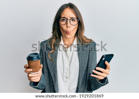 Young brunette woman using smartphone and drinking a cup of coffee making fish face with mouth and squinting eyes, crazy and comical. 