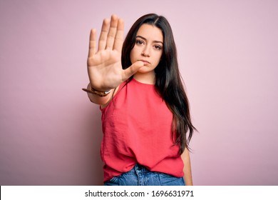 Young brunette woman wearing casual summer shirt over pink isolated background doing stop sing with palm of the hand. Warning expression with negative and serious gesture on the face.