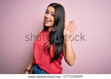 Young brunette woman wearing casual summer shirt over pink isolated background Waiving saying hello happy and smiling, friendly welcome gesture