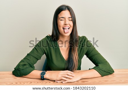 Young brunette woman wearing casual clothes sitting on the table sticking tongue out happy with funny expression. emotion concept. 
