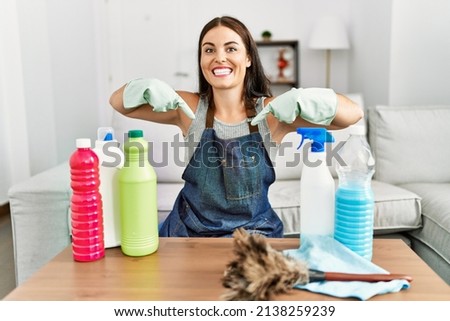 Young brunette woman wearing cleaner apron and gloves cleaning at home looking confident with smile on face, pointing oneself with fingers proud and happy. 