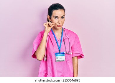 Young brunette woman wearing doctor uniform and stethoscope mouth and lips shut as zip with fingers. secret and silent, taboo talking 