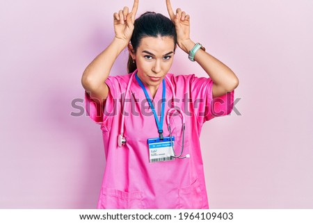 Young brunette woman wearing doctor uniform and stethoscope doing funny gesture with finger over head as bull horns 