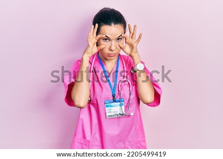 Young brunette woman wearing doctor uniform and stethoscope trying to open eyes with fingers, sleepy and tired for morning fatigue 