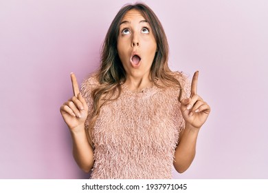 Young brunette woman wearing fashion shirt amazed and surprised looking up and pointing with fingers and raised arms. 