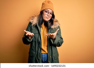 Young Brunette Woman Wearing Glasses And Winter Coat With Hat Over Yellow Isolated Background Clueless And Confused Expression With Arms And Hands Raised. Doubt Concept.