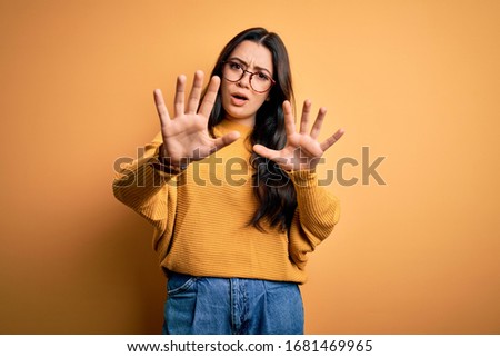 Young brunette woman wearing glasses and casual sweater over yellow isolated background doing stop gesture with hands palms, angry and frustration expression