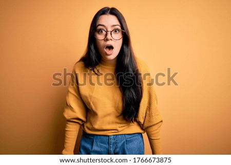 Young brunette woman wearing glasses and casual sweater over yellow isolated background afraid and shocked with surprise and amazed expression, fear and excited face.