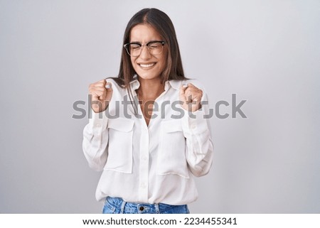 Young brunette woman wearing glasses excited for success with arms raised and eyes closed celebrating victory smiling. winner concept. 