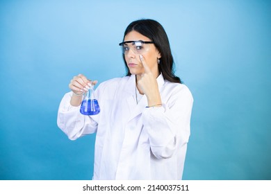 Young Brunette Woman Wearing Scientist Uniform Holding Test Tube Over Isolated Blue Background Pointing To The Eye Watching You Gesture, Suspicious Expression