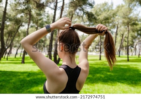 Young brunette woman wearing sportive clothes on city park, outdoors tying her hair in a ponytail while doing some workout.