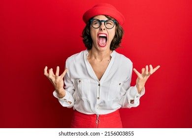 Young brunette woman wearing stewardess style and glasses crazy and mad shouting and yelling with aggressive expression and arms raised. frustration concept. 