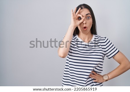 Young brunette woman wearing striped t shirt doing ok gesture shocked with surprised face, eye looking through fingers. unbelieving expression. 