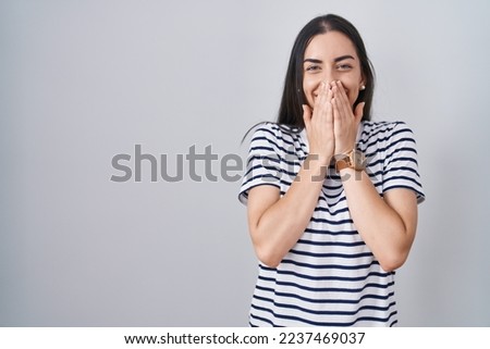 Young brunette woman wearing striped t shirt laughing and embarrassed giggle covering mouth with hands, gossip and scandal concept 