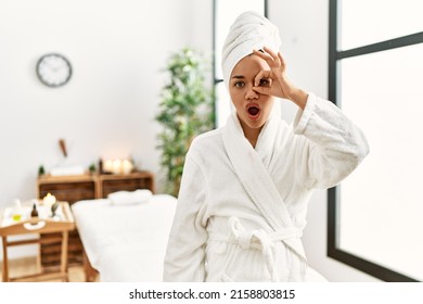 Young brunette woman wearing towel and bathrobe standing at beauty center doing ok gesture shocked with surprised face, eye looking through fingers. unbelieving expression. 