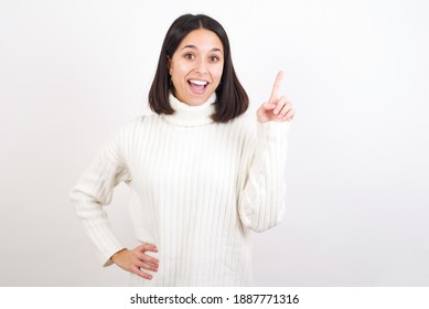 Young Brunette Woman Wearing White Knitted Sweater Against White Background Holding Finger Up Having Idea And Posing