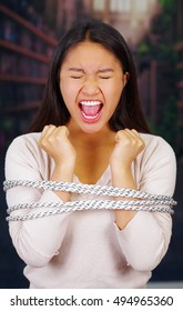 Young Brunette Woman Wearing White Sweater Tied Up With Rope Around Upper Body, Interacting Breaking Free And Screaming, Facing Camera, Hostage Concept