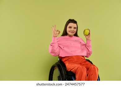 A young brunette woman in a wheelchair sits in a studio setting, wearing casual attire. She holds a green apple in one hand and makes an OK sign with the other. - Powered by Shutterstock