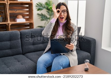 Young brunette woman working at consultation office doing ok gesture shocked with surprised face, eye looking through fingers. unbelieving expression. 