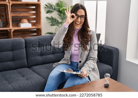 Young brunette woman working at consultation office doing ok gesture with hand smiling, eye looking through fingers with happy face. 