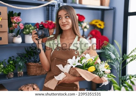 Young brunette woman working at florist shop holding camera smiling looking to the side and staring away thinking. 