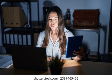 Young brunette woman working at the office at night puffing cheeks with funny face. mouth inflated with air, crazy expression. 