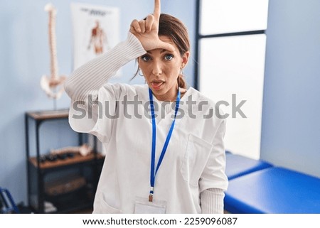 Young brunette woman working at pain recovery clinic making fun of people with fingers on forehead doing loser gesture mocking and insulting. 