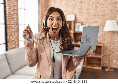Young brunette woman working as real state agent holding keys of new home angry and mad screaming frustrated and furious, shouting with anger. rage and aggressive concept. 