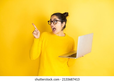 Young Brunette Woman Working Using Computer Laptop Over Yellow Background Amazed And Pointing With Hand And Finger To The Side