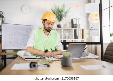 Young builder  engineer in reflective vest uniform   hard hat desk in office background large TV screen and   browsing in laptop   looking through papers sketches 