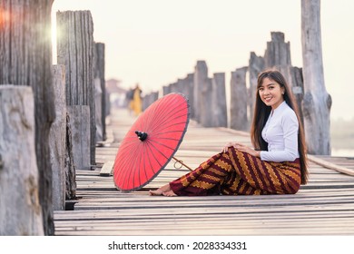 A young Burmese woman wearing traditional clothes with red umbrella at  U Bein teak bridge on Taungthaman lake in Mandalay Division. Myanmar. - Shutterstock ID 2028334331