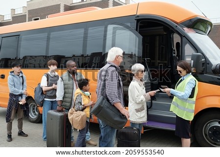 Young bus conductor checking tickets by open door of vehicle with line if passengers in front