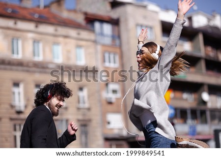 Young business colleagues dancing together with headphones after work. Happy friends jumping and fooling around outdoors.