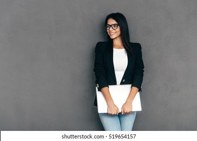 Young Business Expert. Attractive Young Woman In Smart Casual Wear And Eyeglasses Carrying Laptop And Smiling While Standing Against Grey Background