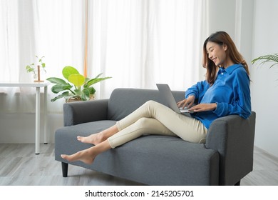 Junge freiberufliche Asiatin, die auf Laptops arbeitet und soziale Medien prüft, während sie auf dem Sofa liegen, wenn sie sich im Wohnzimmer zu Hause entspannen.