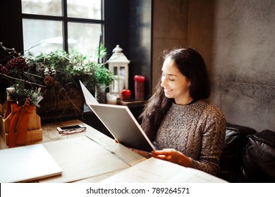 Young Business Girl Studying The Menu In Restaurant Decorated With Christmas Decor.sits Near The Window On Cloudy Winter Day At Wooden Table.Dressed In Warm Gray Sweater.On The Table,phone And Glasses